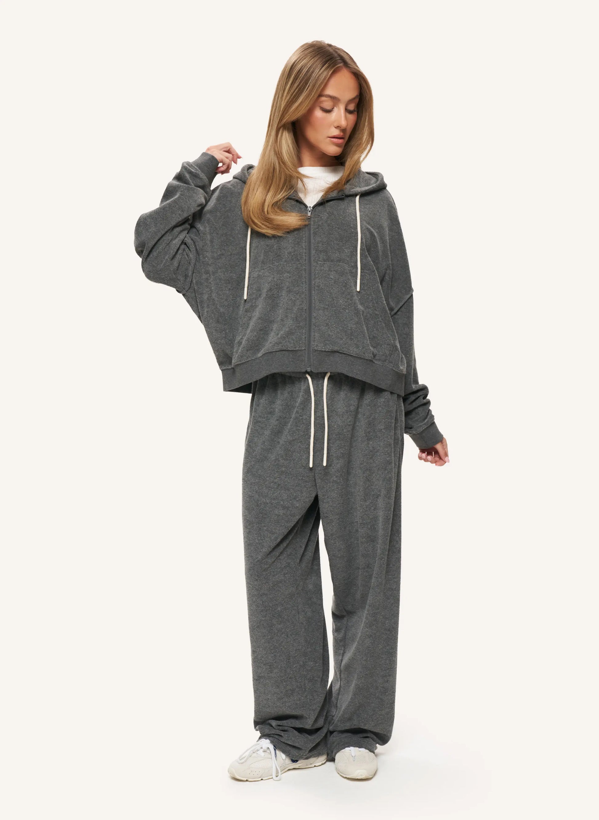 A woman modeling a dark gray two-piece loungewear set consisting of a cropped zip-up hoodie and matching sweatpants, styled with a white t-shirt and white sneakers against a light off-white background.