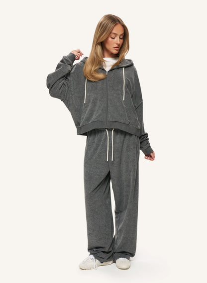 A woman modeling a dark gray two-piece loungewear set consisting of a cropped zip-up hoodie and matching sweatpants, styled with a white t-shirt and white sneakers against a light off-white background.