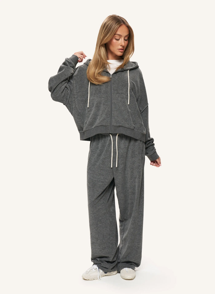 A woman modeling a dark gray two-piece loungewear set consisting of a cropped zip-up hoodie and matching sweatpants, styled with a white t-shirt and white sneakers against a light off-white background.