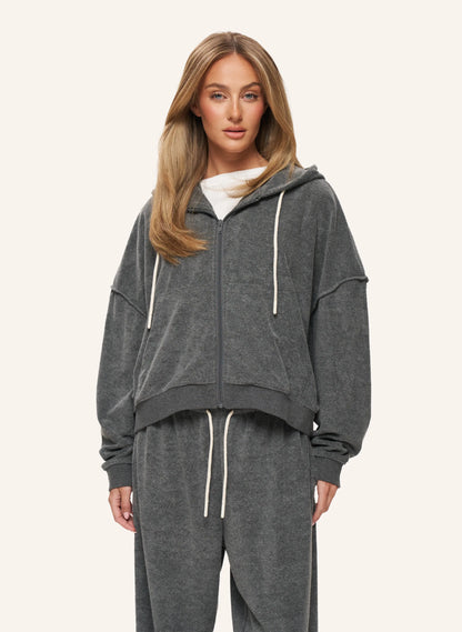 A woman modeling a dark gray heather zip-up hoodie and matching jogger pants set with white drawstrings, photographed against a solid off-white background.