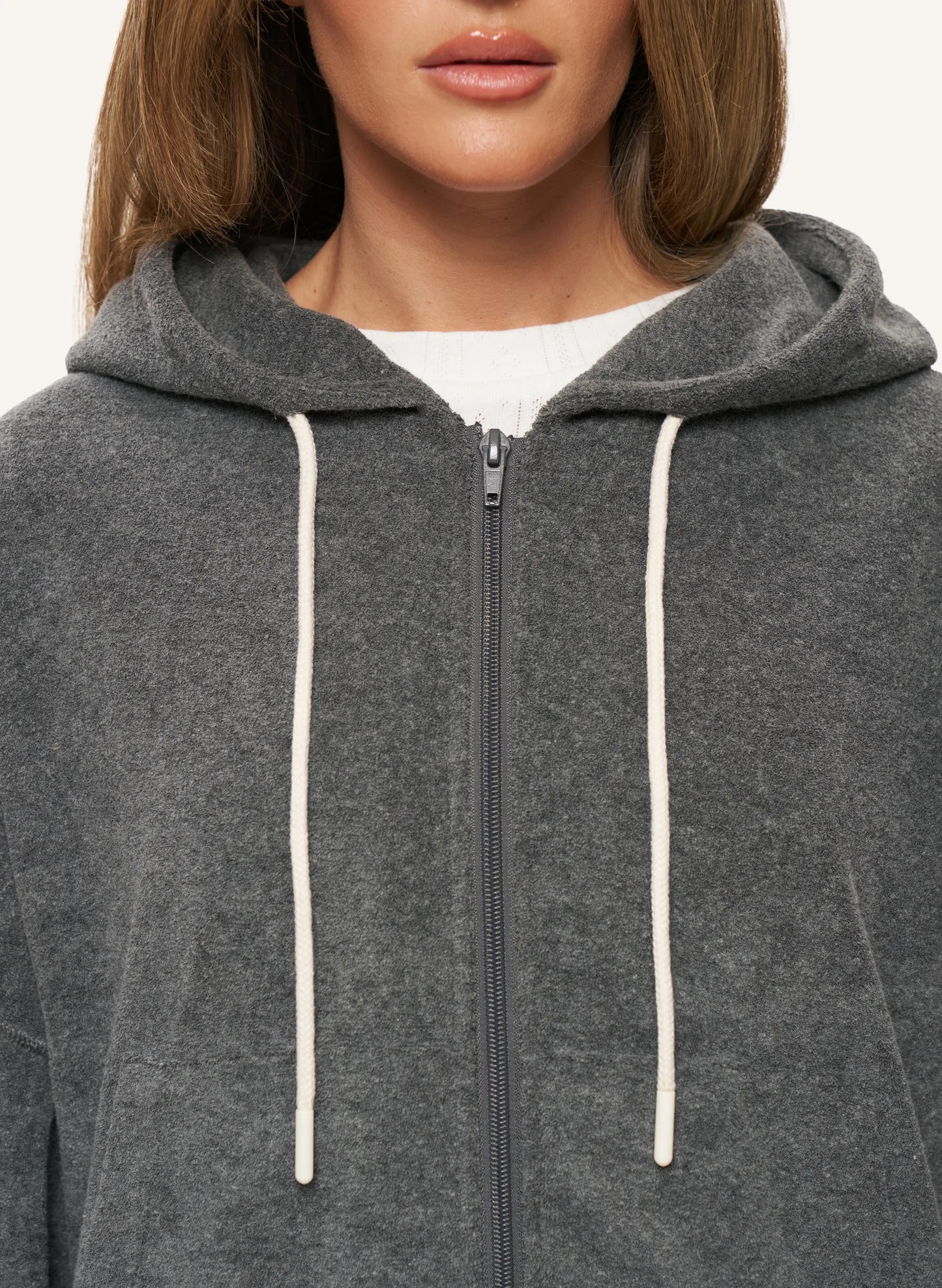 A close-up of a model wearing a dark gray zip-up hoodie with cream-colored drawstrings over a white ribbed top.