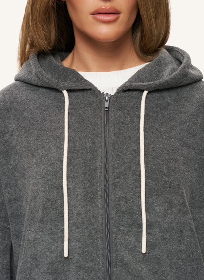 A close-up of a model wearing a dark gray zip-up hoodie with cream-colored drawstrings over a white ribbed top.