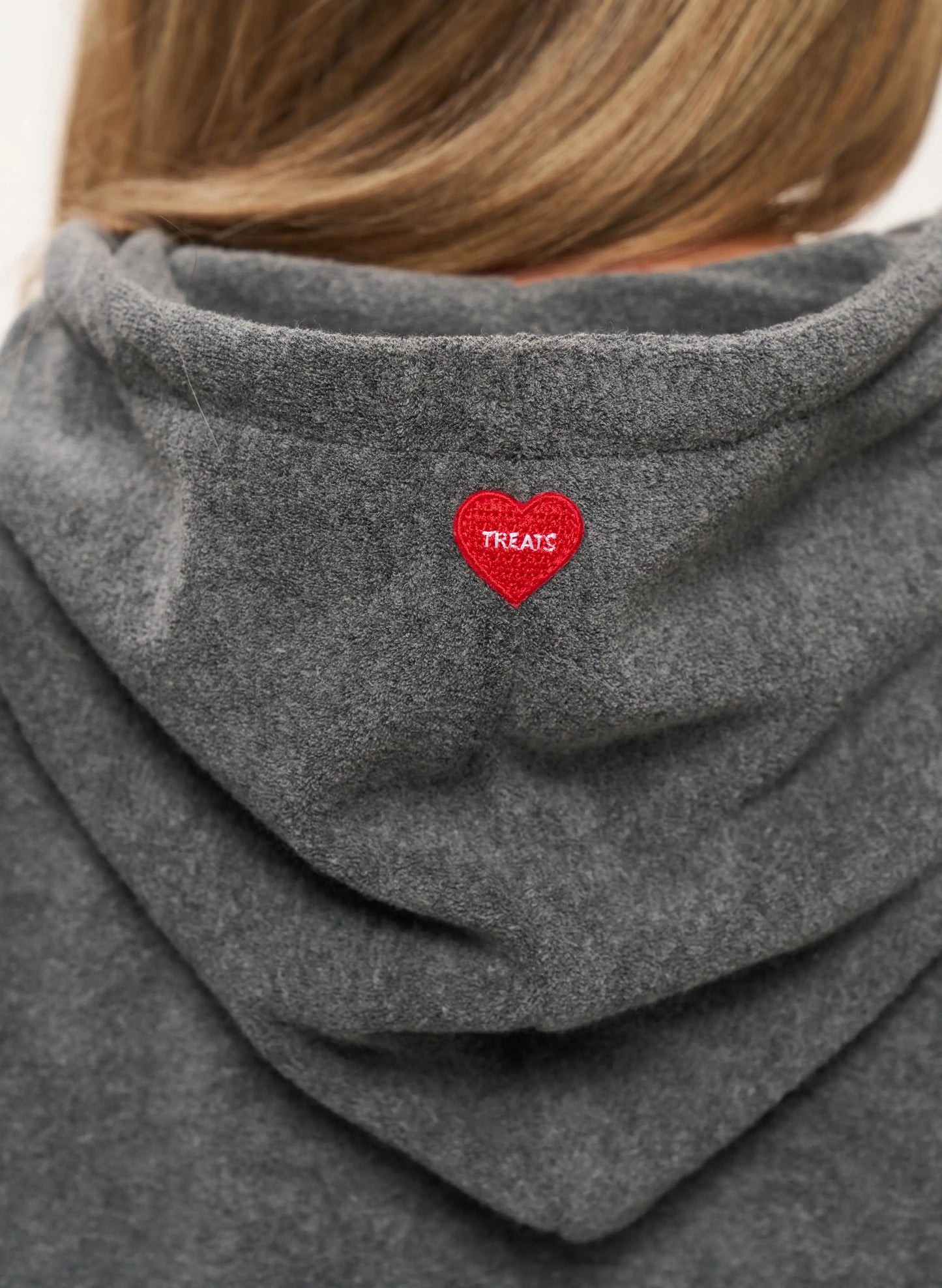 Close-up of the back of a person wearing a heather gray hooded sweatshirt, with a small red heart patch on the back of the neck that reads 'TREATS'.