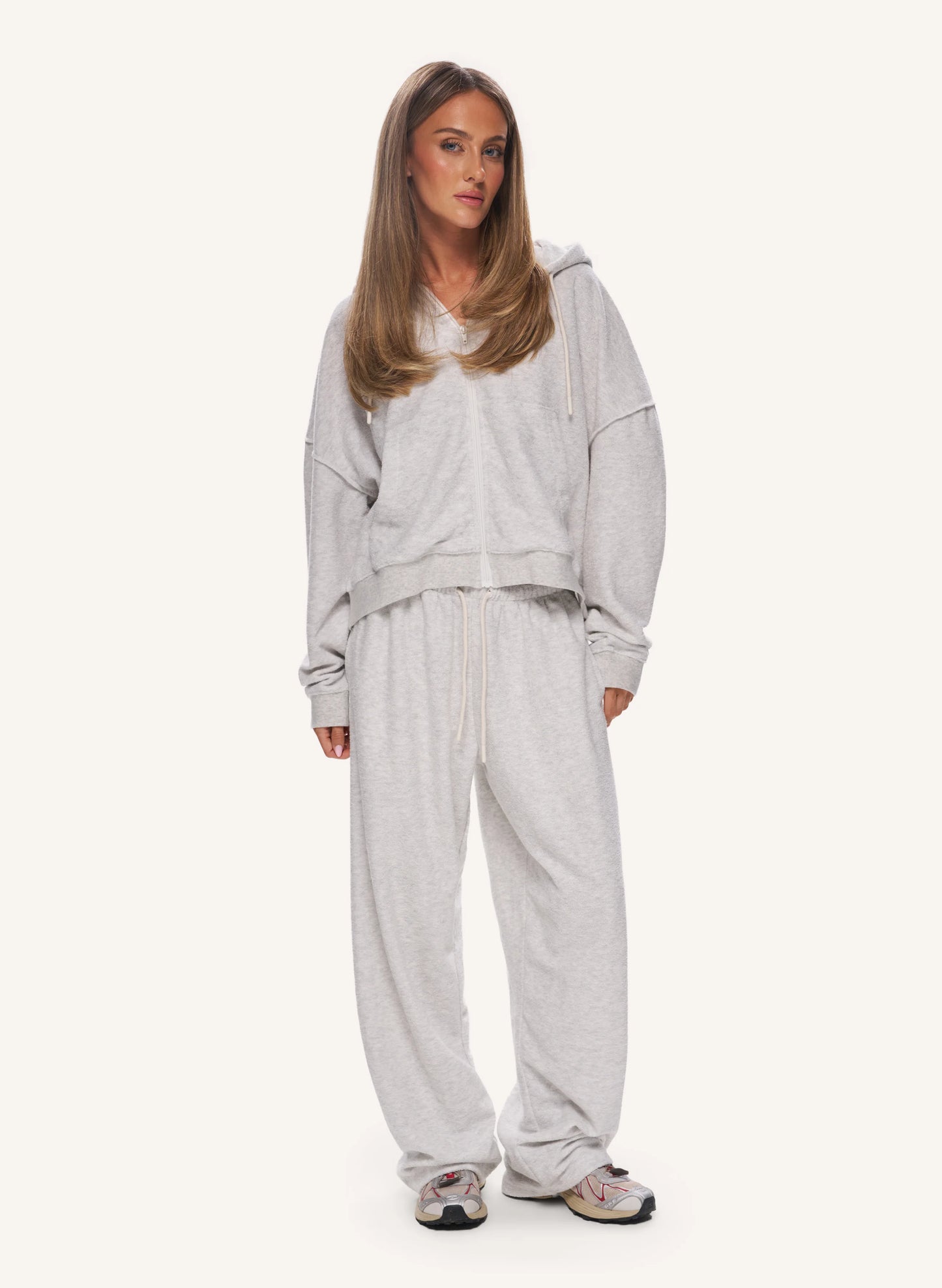 A woman modeling a light grey heathered lounge set consisting of a cropped hoodie and matching sweatpants, paired with grey sneakers, against a solid off-white background.