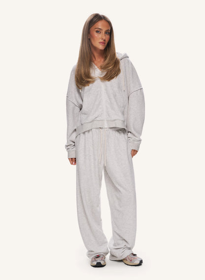 A woman modeling a light grey heathered lounge set consisting of a cropped hoodie and matching sweatpants, paired with grey sneakers, against a solid off-white background.