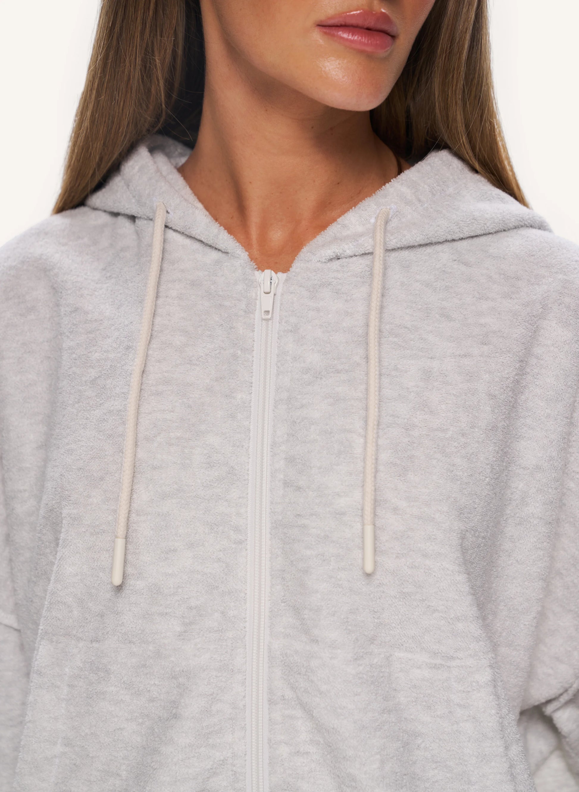 A close-up of a model wearing a light heather gray zip-up hoodie with cream-colored drawstrings against a plain white background.