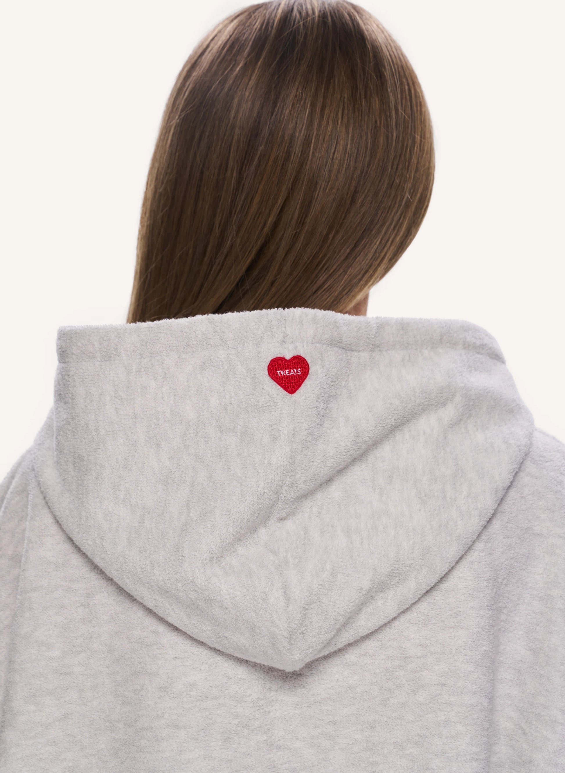 Back view of a person wearing a light heather gray hoodie, featuring a small red embroidered heart with white text on the back of the neck.