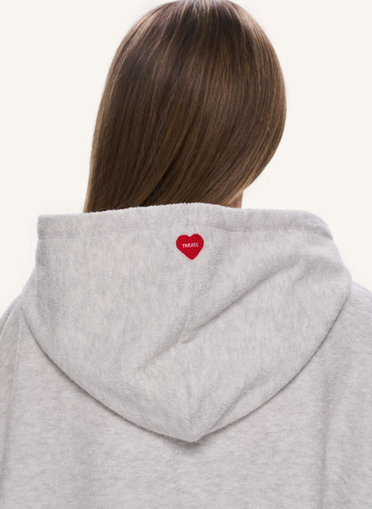 Back view of a person wearing a light heather gray hoodie, featuring a small red embroidered heart with white text on the back of the neck.
