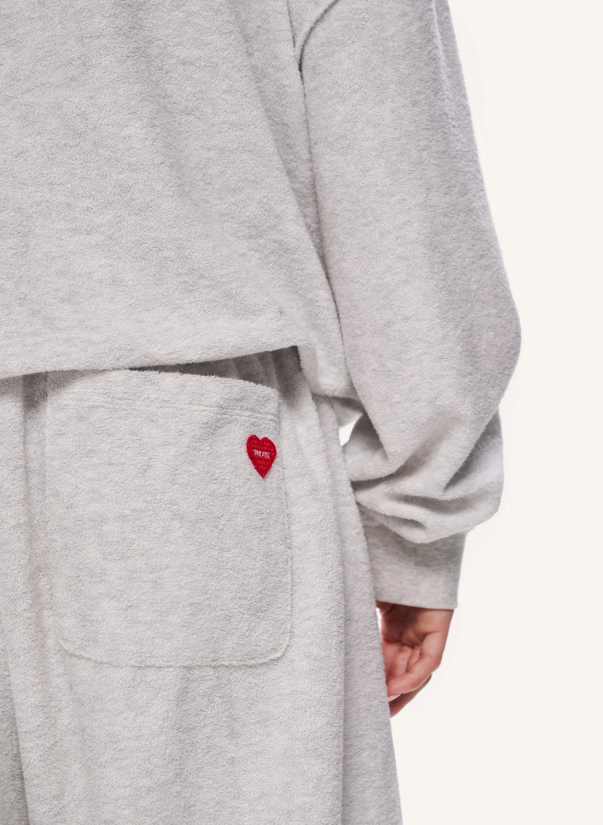 Close-up of a person wearing a light heather grey sweatshirt or loungewear set, showing a front pocket with a small red embroidered heart logo and part of the wearer's hand.