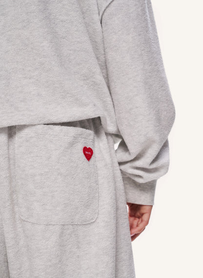 Close-up of a person wearing a light heather grey sweatshirt or loungewear set, showing a front pocket with a small red embroidered heart logo and part of the wearer's hand.