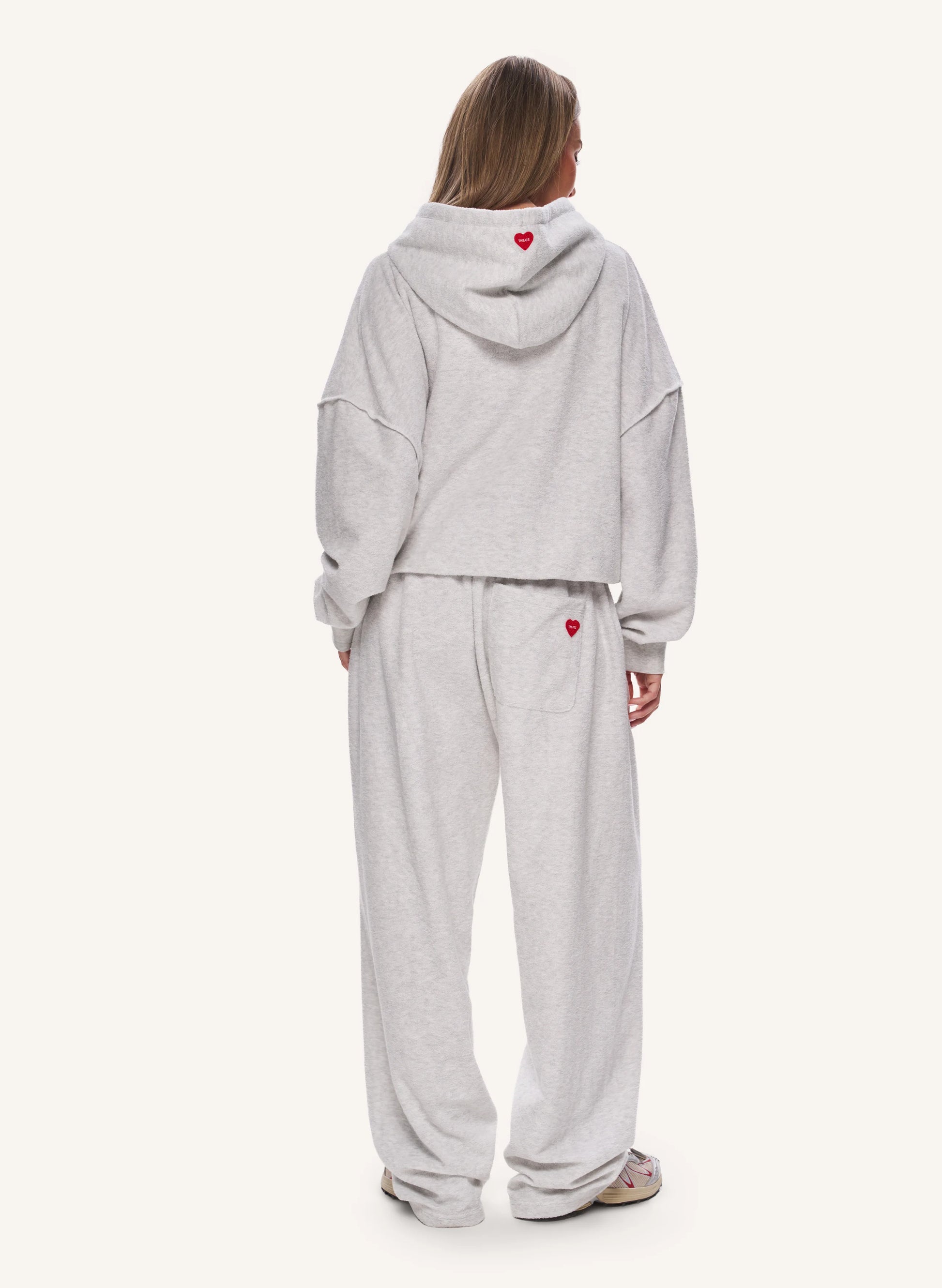 A model viewed from the back wearing a light grey heather cropped hoodie and matching sweatpants, both featuring a small red heart detail.