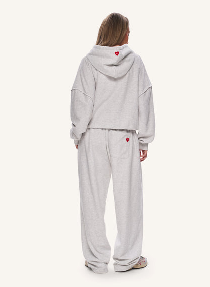 A model viewed from the back wearing a light grey heather cropped hoodie and matching sweatpants, both featuring a small red heart detail.