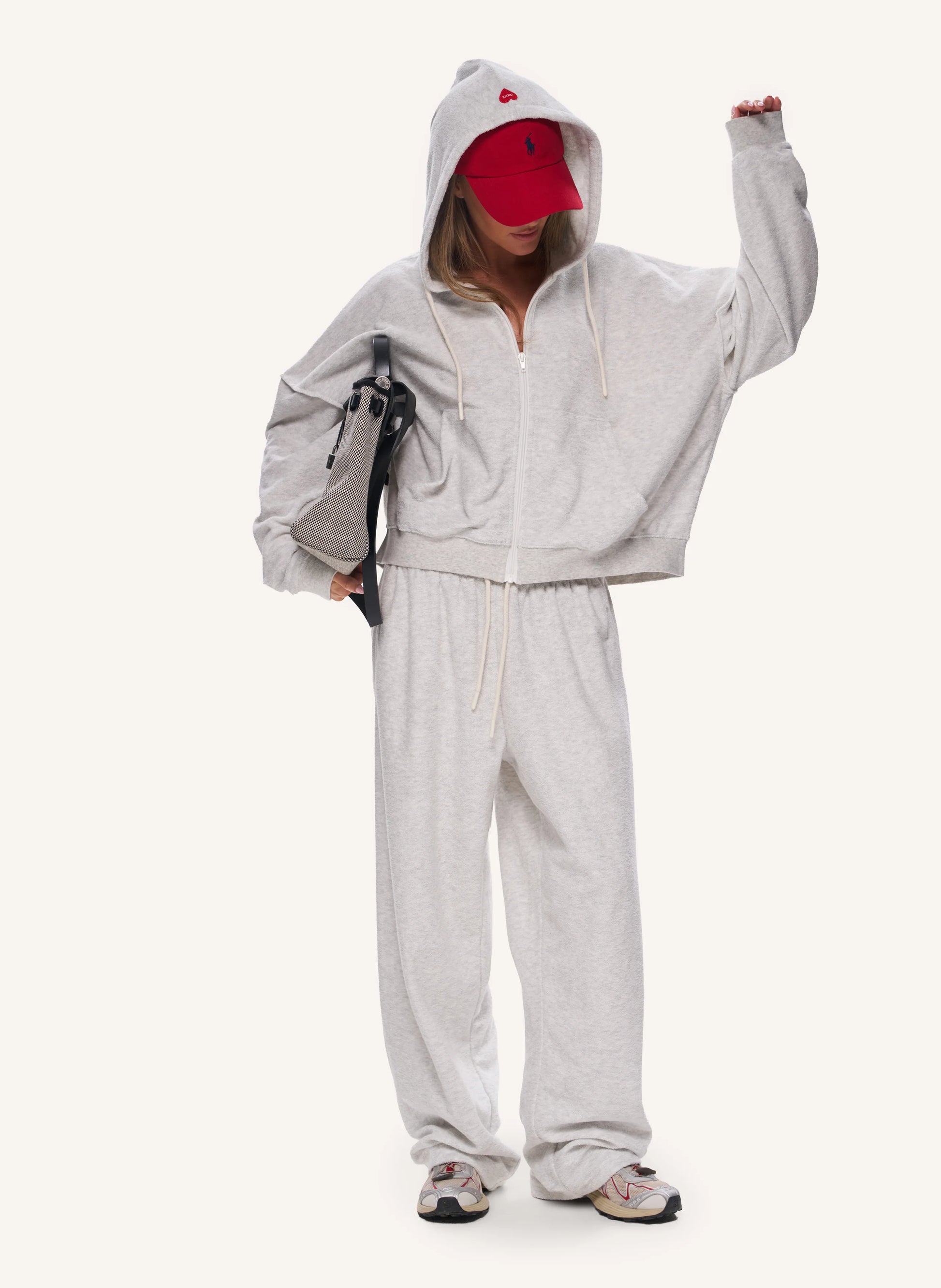 A model wearing a light gray heathered zip-up hoodie and matching sweatpants, styled with a red baseball cap, a grey shoulder bag, and sneakers, set against a plain off-white background.