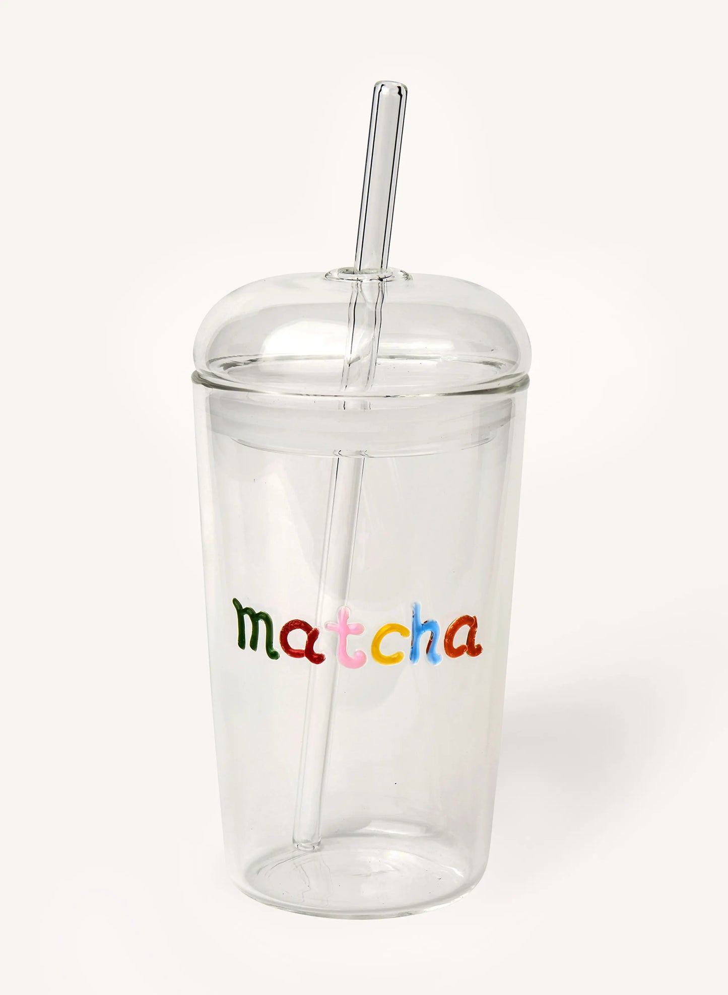 A clear glass tumbler with a matching glass lid and straw, featuring the word 'matcha' printed in colorful lowercase letters on the front, set against a plain off-white background.