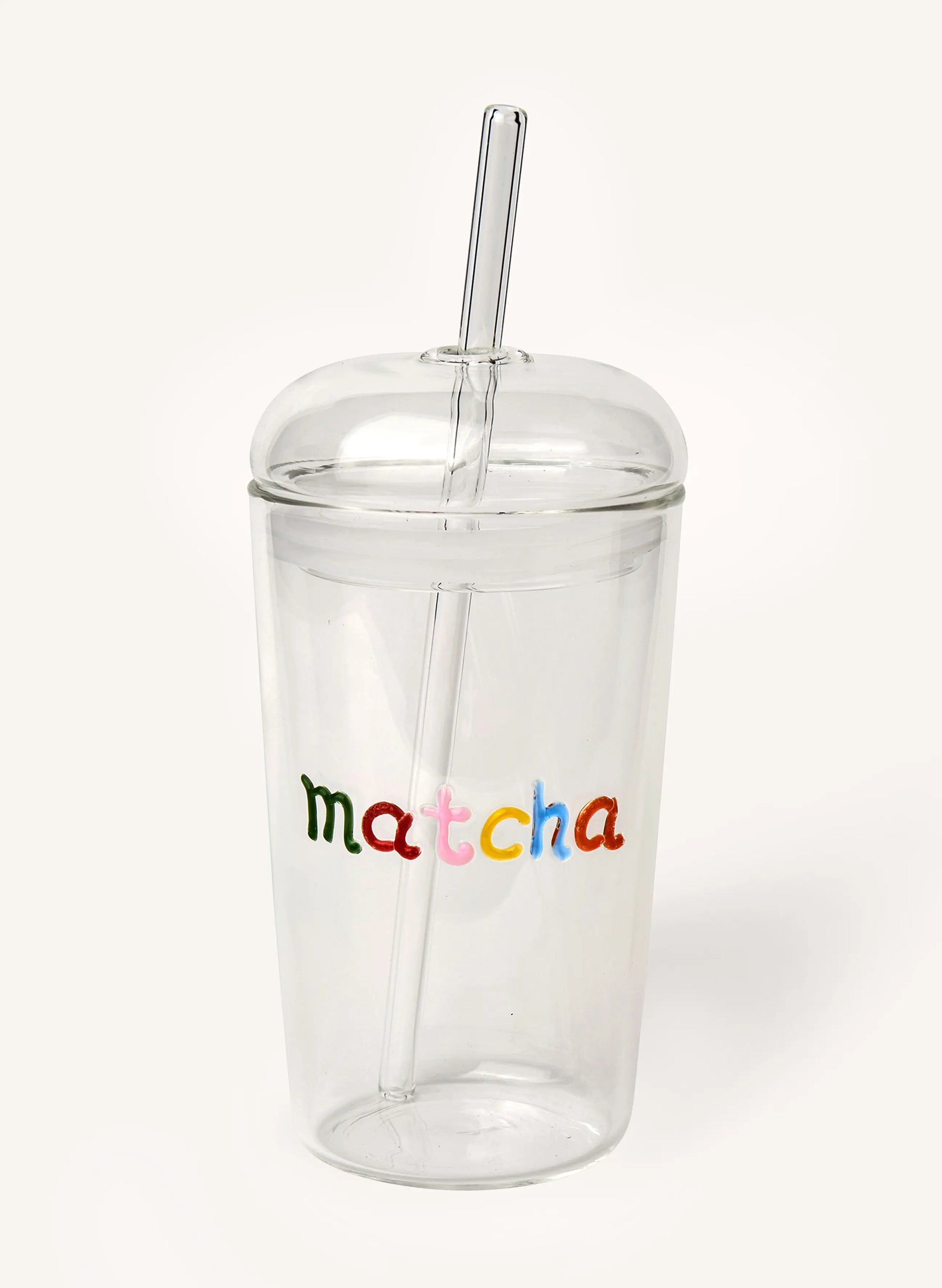 A clear glass tumbler with a matching glass lid and straw, featuring the word 'matcha' printed in colorful lowercase letters on the front, set against a plain off-white background.