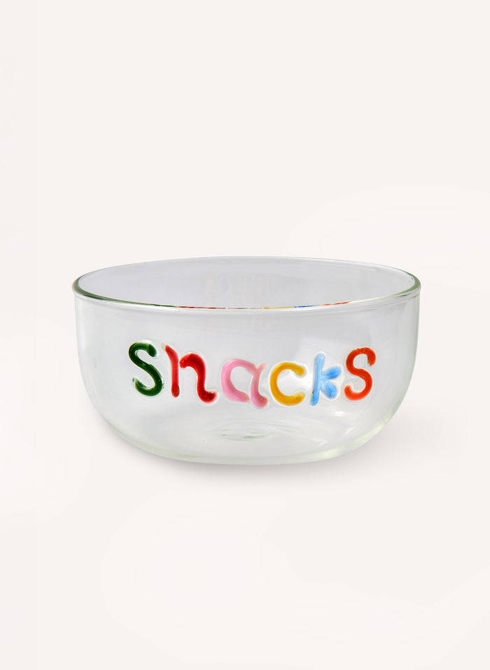A clear glass bowl with the word 'Snacks' written across the front in colorful, playful letters, set against a plain off-white background.