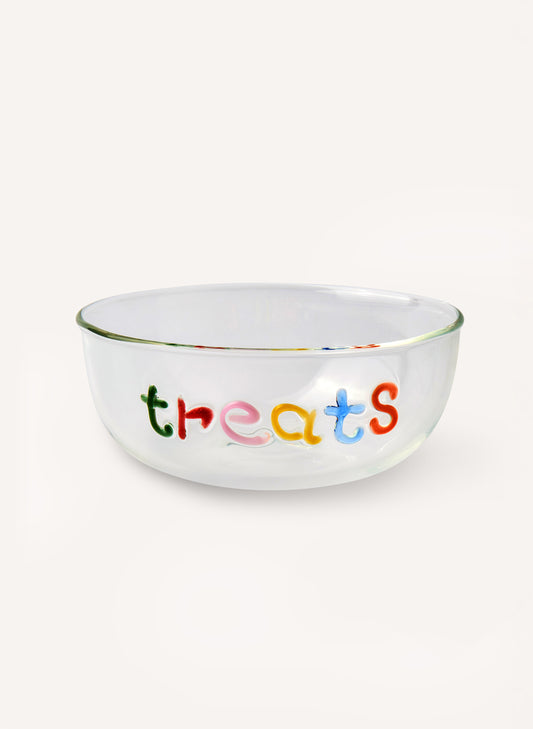 A clear glass bowl with the word 'treats' written across the front in colorful, playful letters, set against a plain off-white background.