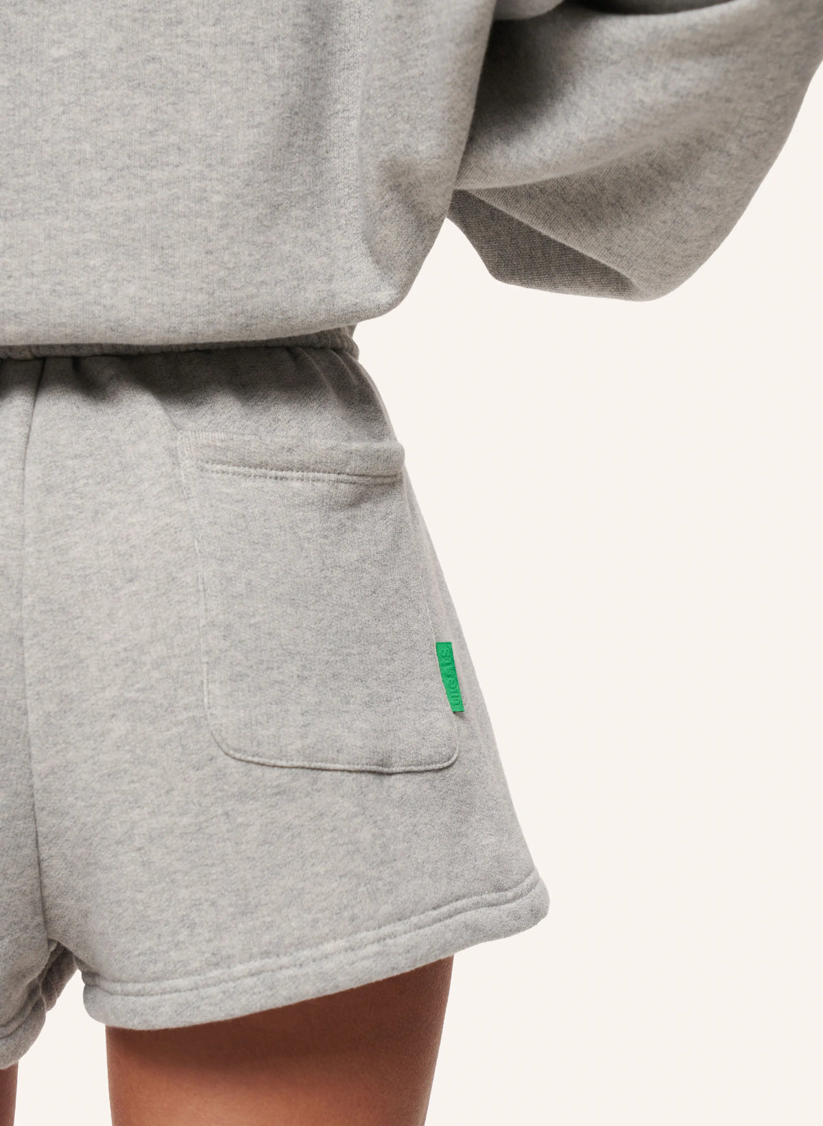 Close-up of a person wearing light heather gray sweat shorts with a side pocket and a small green tag, paired with a matching gray sweatshirt, against a plain off-white background.