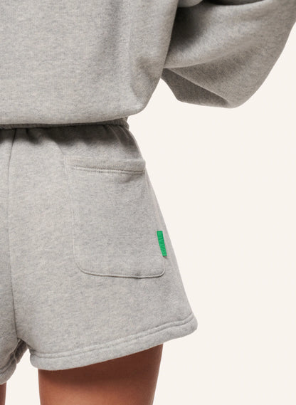 Close-up of a person wearing light heather gray sweat shorts with a side pocket and a small green tag, paired with a matching gray sweatshirt, against a plain off-white background.