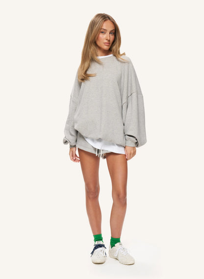 A woman modeling an oversized light gray crewneck sweatshirt, styled with a white t-shirt underneath, white denim shorts, green socks, and white sneakers, against a solid off-white background.