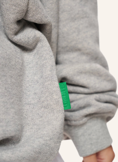 Close-up of a light gray heathered garment, likely a sweater or hoodie sleeve, with a small bright green fabric tag showing the brand name 'TREATS'.