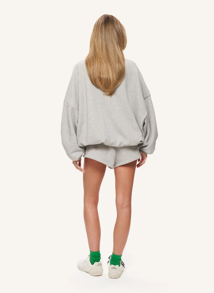 A woman viewed from behind wearing an oversized light gray sweatshirt and matching shorts, with bright green socks and white sneakers, against a plain off-white studio background.