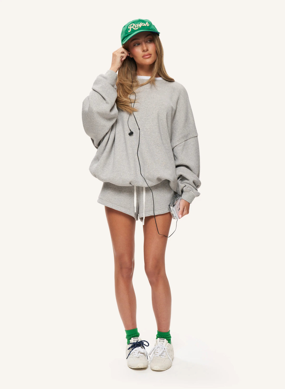 A woman modeling an oversized light gray sweatshirt and matching shorts set, styled with a green baseball cap, green socks, and white sneakers against a plain light background.