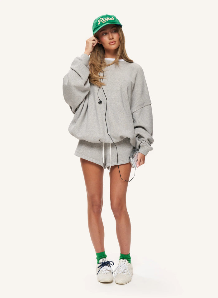 A woman modeling an oversized light gray sweatshirt and matching shorts set, styled with a green baseball cap, green socks, and white sneakers against a plain light background.