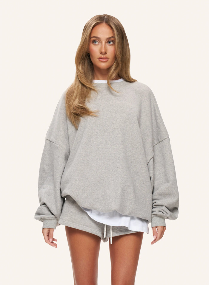 A woman modeling an oversized light gray heather sweatshirt with voluminous long sleeves and a relaxed fit, paired with matching gray shorts and a white undershirt, against a solid off-white background.