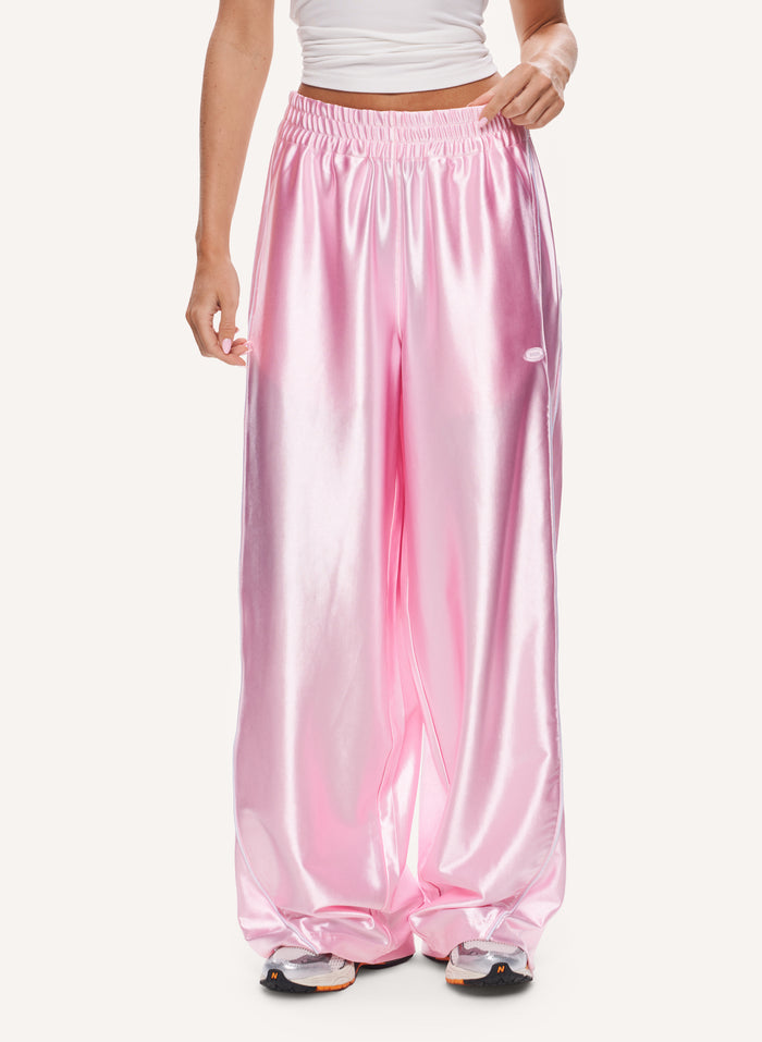 A model wearing shiny light pink wide-leg pants with an elastic waistband, paired with a white crop top and sneakers, against a plain off-white background.