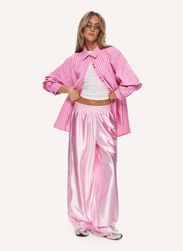 A woman modeling a pink striped button-up shirt and shiny pink satin pants against a plain light background.