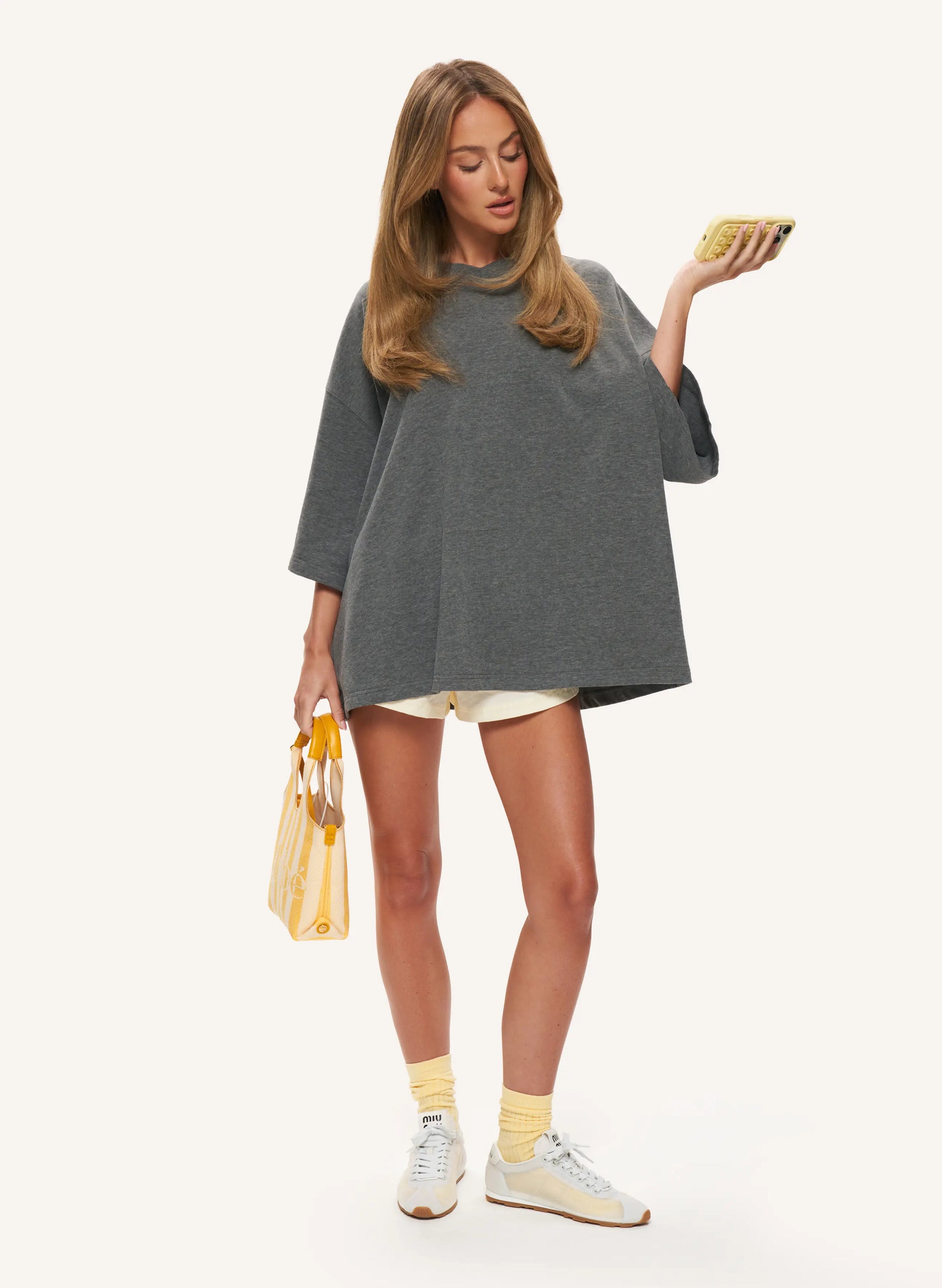 A woman modelling an oversized dark grey  top with elbow length sleeves, paired with light shorts, yellow socks, white sneakers, and holding a yellow patterned handbag against a plain off-white background.