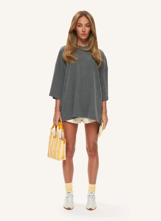 A woman stands against a plain light background wearing an oversized dark gray top, lemmon shorts, yellow socks, white sneakers, and holding a yellow striped tote bag.