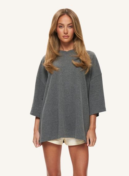 A woman modelling a loose-fitting, oversized gray top with wide three-quarter sleeves, paired with light beige shorts, against a solid off-white background.