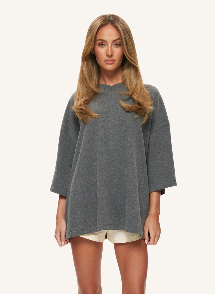 A woman modelling a loose-fitting, oversized gray top with wide three-quarter sleeves, paired with light beige shorts, against a solid off-white background.