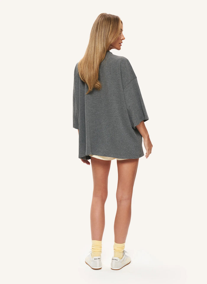 A woman shown from the back wearing an oversized dark grey top, lemon shorts, yellow socks, and white sneakers against a plain off-white studio background