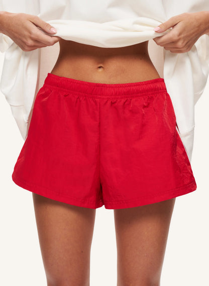 A close-up of a model wearing bright red athletic shorts, holding up a white top to show the fit. The background is a clean, solid off-white studio setting.