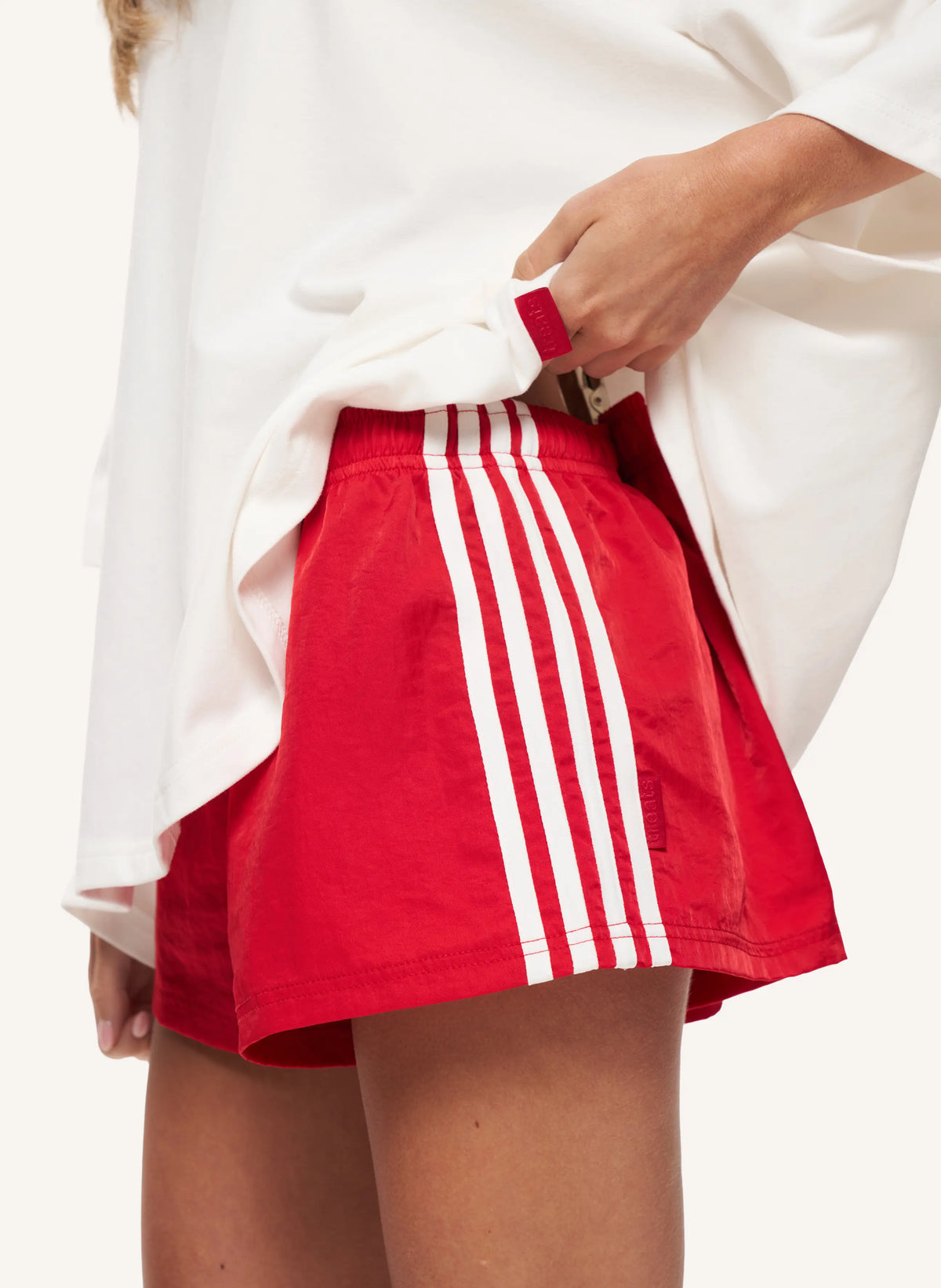 A close-up of a person wearing bright red athletic shorts with three white vertical stripes down the side, paired with an oversized white top. The person is pulling the hem of the shirt slightly to show the fit of the shorts.