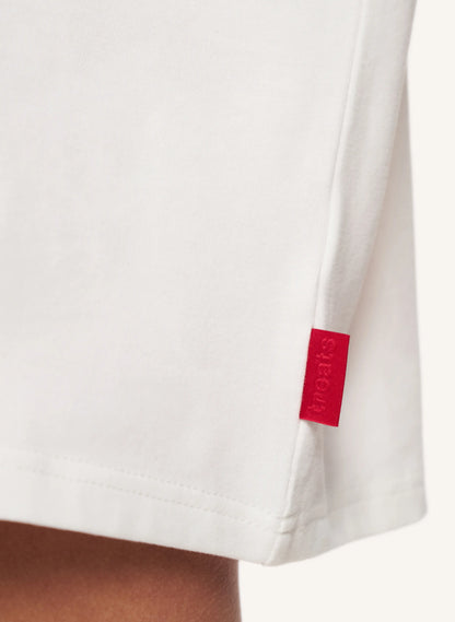 Close-up of a white short-sleeve garment with a small red fabric brand tab on the sleeve, worn on a model's arm against a light neutral background.
