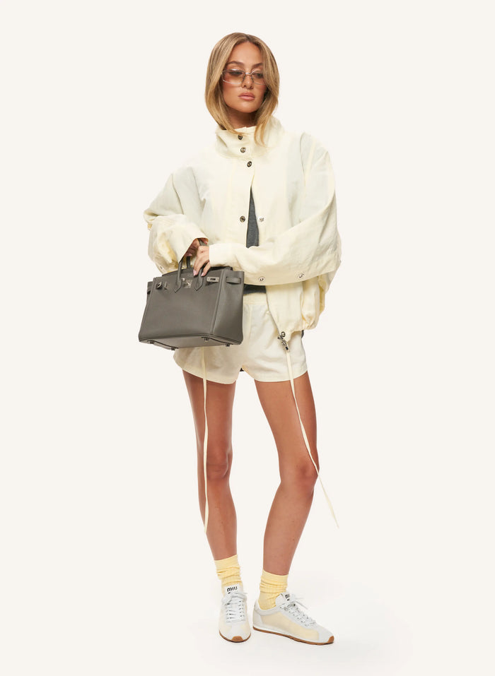 A model stands against a plain off-white studio background wearing a cream hooded jacket, matching cream shorts, yellow socks, white sneakers, and holding a dark gray structured handbag.