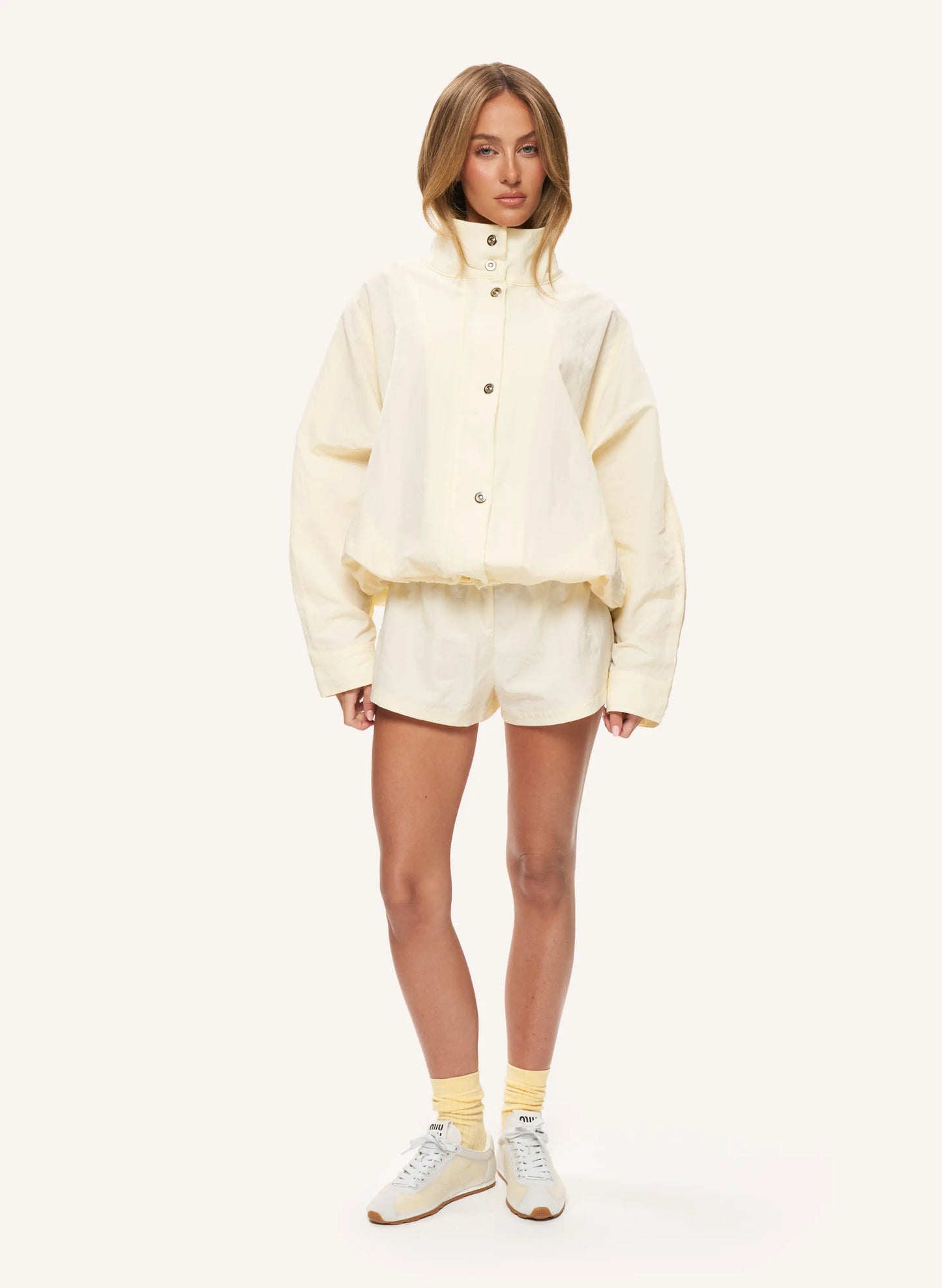 A model stands against a plain off-white studio background wearing a cream-colored cropped button-front jacket with a high collar, matching shorts, yellow socks, and light-colored sneakers.