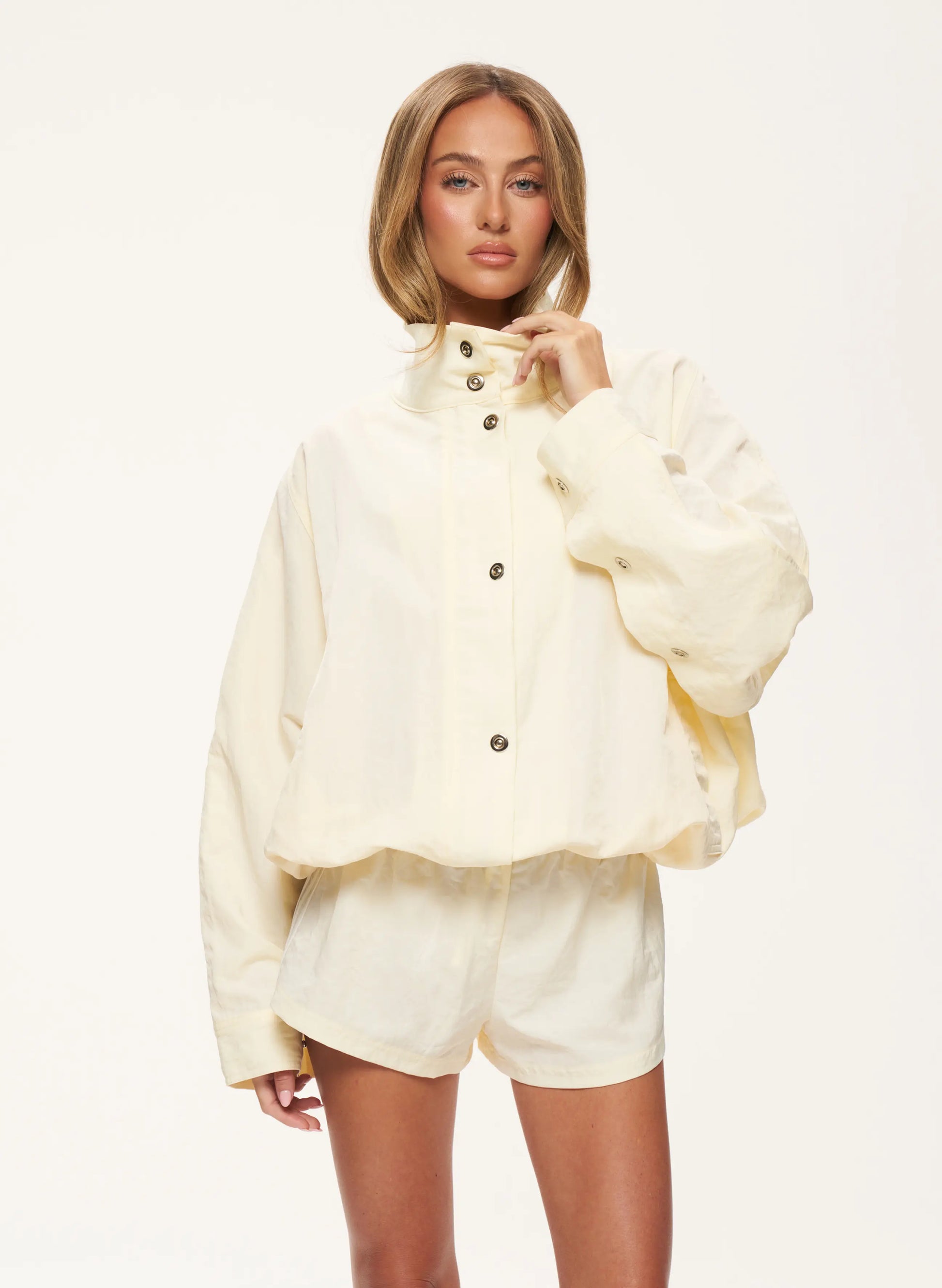 A model wearing a cream-colored cropped jacket with a high collar and matching shorts, set against a plain white studio background.