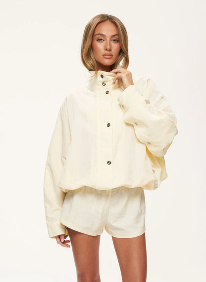 A model wearing a cream-colored cropped jacket with a high collar and matching shorts, set against a plain white studio background.