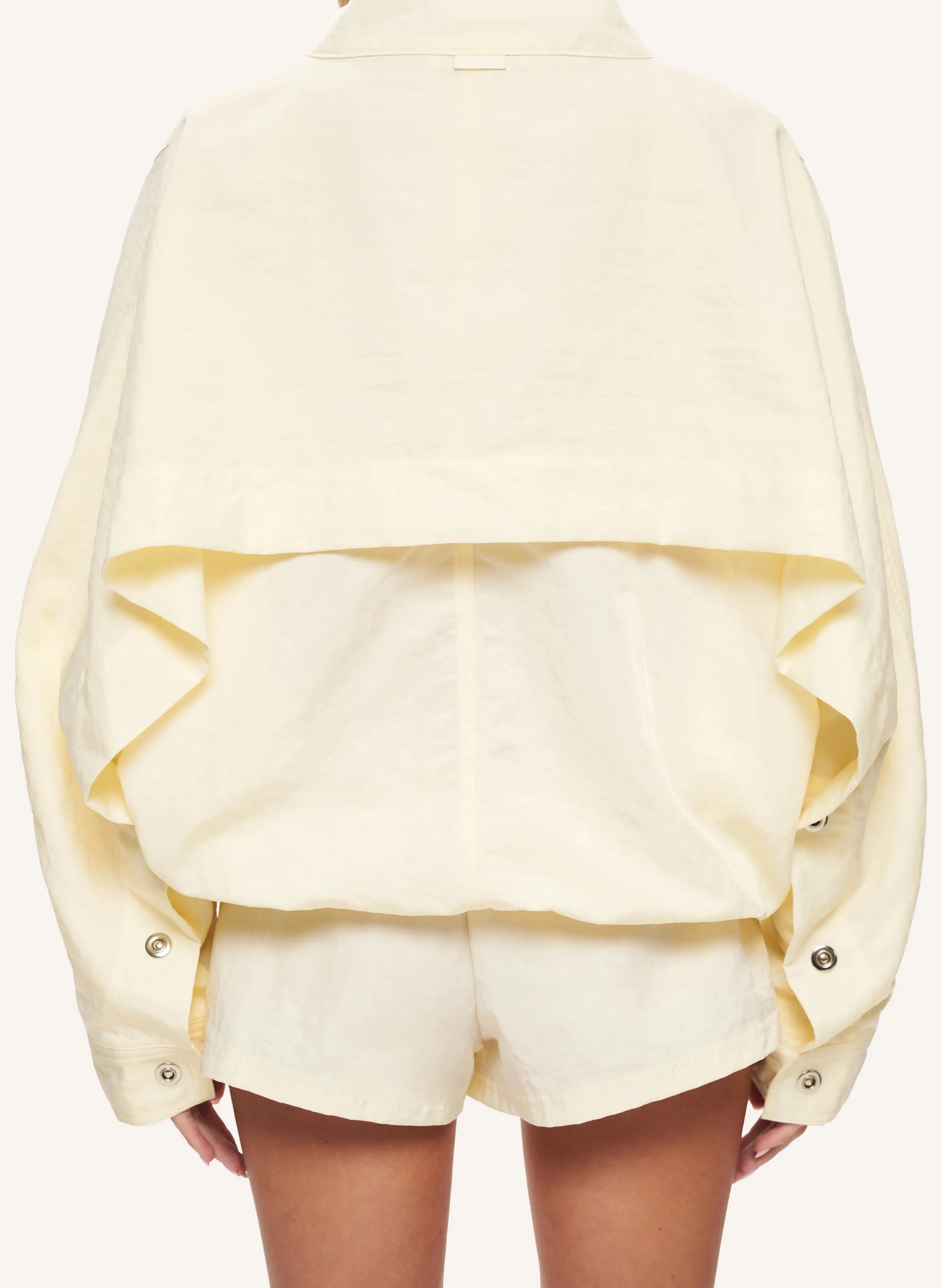 A pale yellow cropped jacket with dramatic ruffles and button details, shown from the back on a model wearing matching shorts against a light cream background.