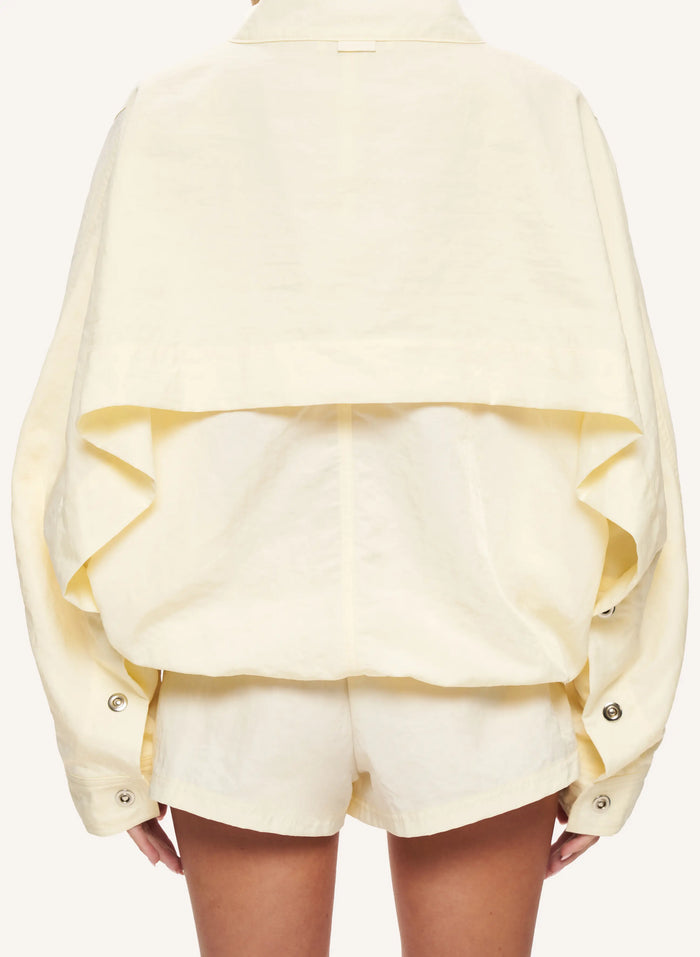 A pale yellow cropped jacket with dramatic ruffles and button details, shown from the back on a model wearing matching shorts against a light cream background.