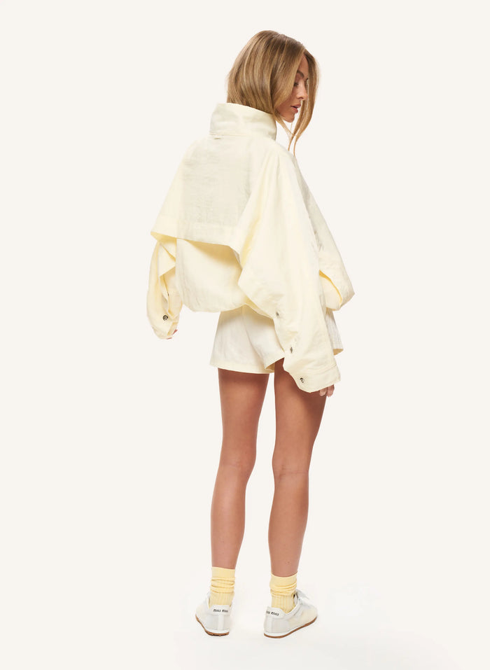 A model shown from the back wearing a cream-colored oversized cropped jacket, matching shorts, yellow socks, and white sneakers against a solid off-white background.