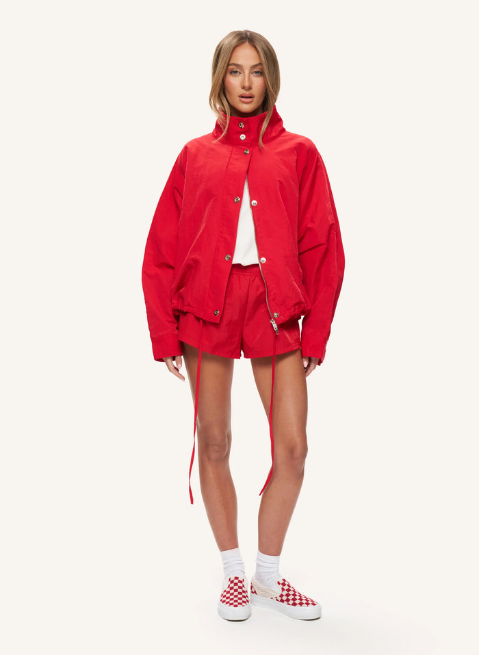A model stands against a light off-white studio background wearing a bright red cropped jacket, matching red shorts, a white top, white socks, and red-and-white checkered sneakers.