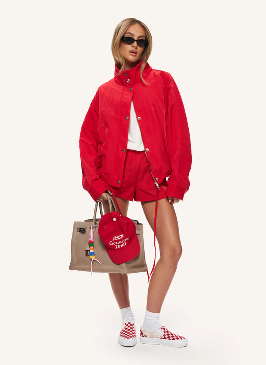 A model stands against a plain light background wearing a bright red matching jacket and shorts set, a white top, red-and-white checkered sneakers, white socks, sunglasses, and carrying a beige tote bag with a red cap.