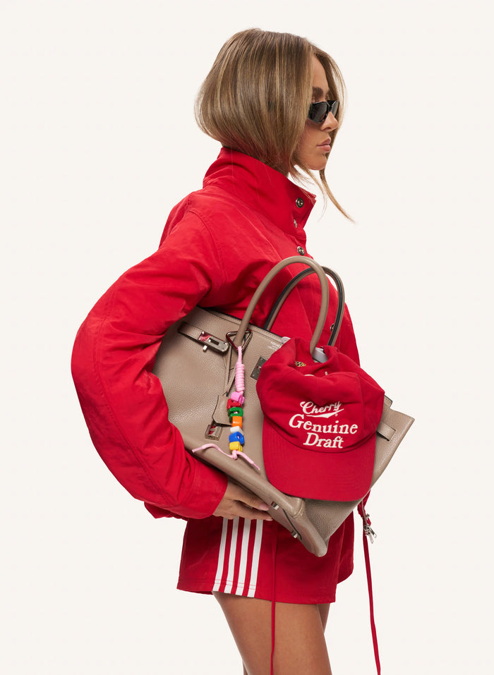 A model in a red tracksuit and sunglasses is shown from the side, carrying a beige leather handbag and wearing a red baseball cap with 'Genuine Draft' embroidered in white. The background is a solid off-white studio setting.