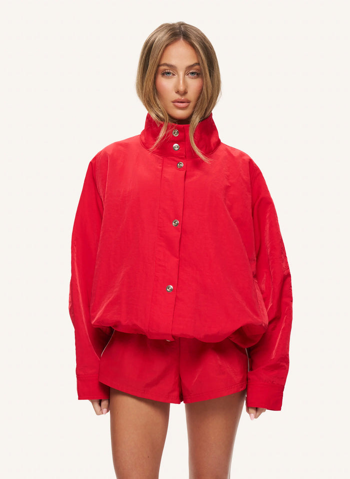 A model wearing a bright red cropped jacket with a high stand-up collar and matching red shorts, photographed against a plain light gray studio background.
